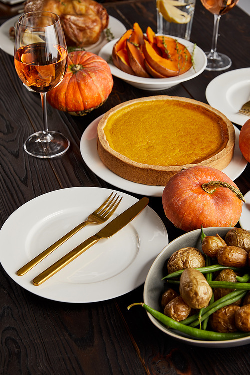 traditional thanksgiving dinner with pumpkin pie, baked vegetables and whole pumpkins near plate with golden fork and knife on dark wooden surface