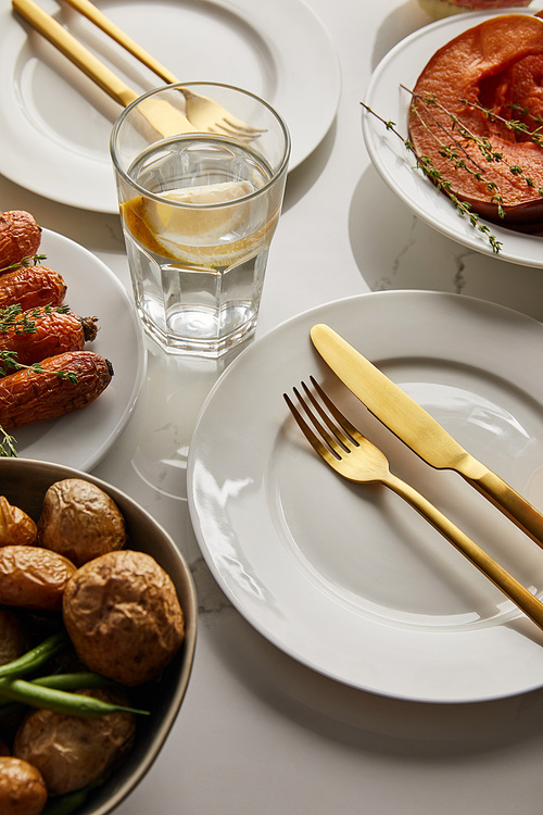 white plates with baked vegetables, golden forks and knives, glass with lemon water on white marble table