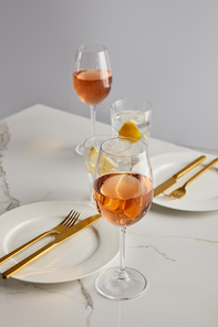 glasses with rose wine and lemon water near white plates with forks and knives on marble table isolated on grey