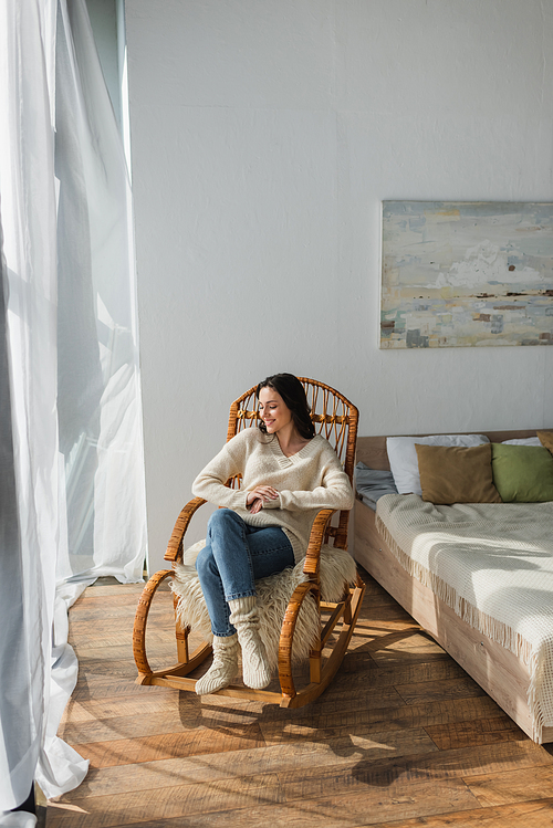 happy woman in warm sweater and knitted socks relaxing in rocking chair in bedroom
