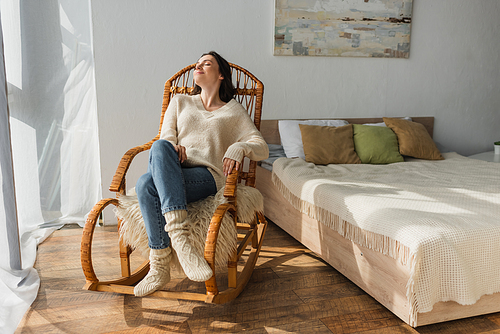 joyful woman in soft sweater and warm socks resting in rocking chair in bedroom