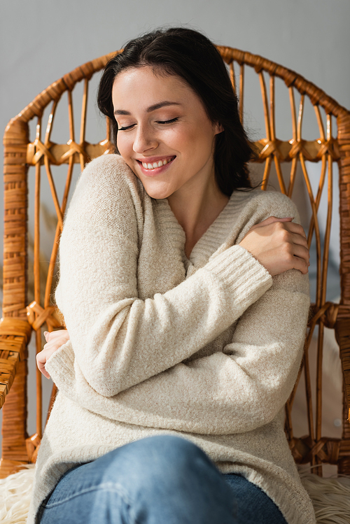 smiling woman in cozy sweater hugging herself in wicker chair