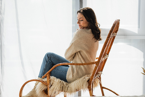 brunette woman in warm sweater and jeans resting in rocking chair at home