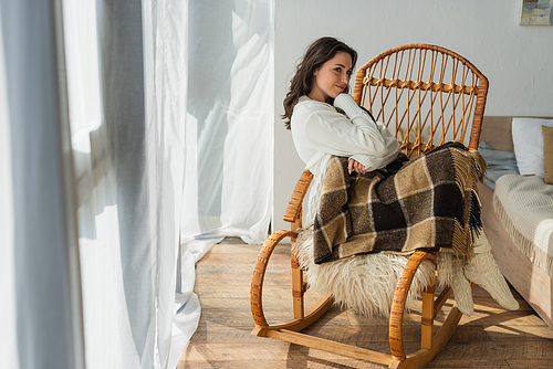 brunette woman smiling and looking away while resting in rocking chair under plaid blanket