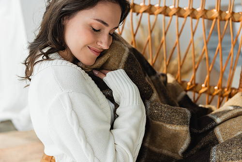 brunette woman sitting in wicker chair under plaid blanker and smiling with closed eyes