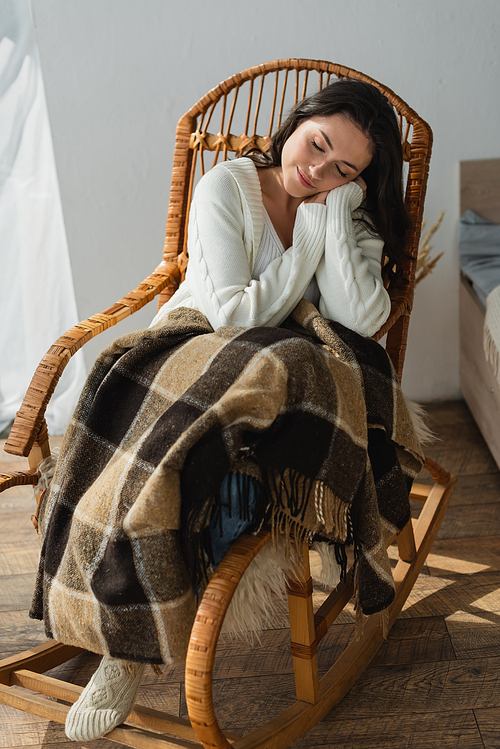 brunette woman in warm cardigan smiling while sleeping in wicker chair under plaid blanket