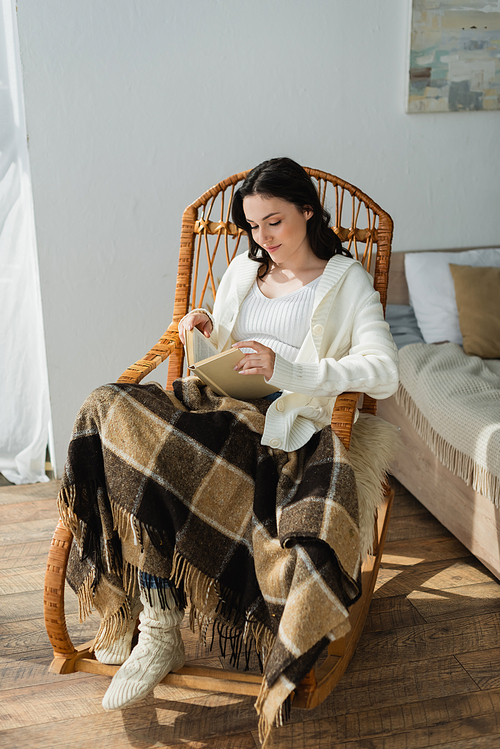 smiling woman sitting in wicker chair under plaid blanket and reading book
