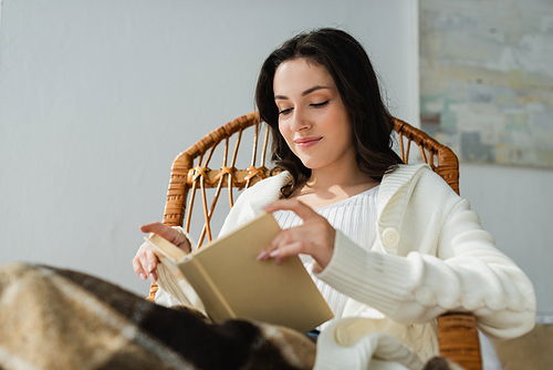 young woman smiling while reading book in wicker chair under checkered blanket