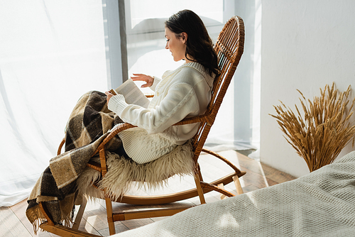 side view of brunette woman reading book in rocking chair under plaid blanket