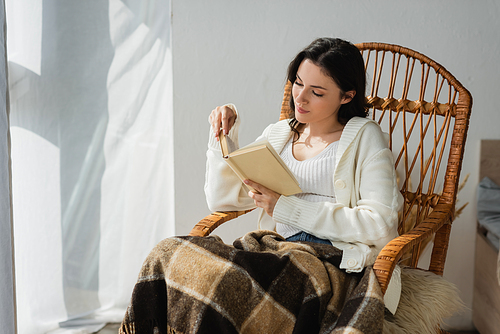 brunette woman in white cardigan reading novel in whicker chair under warm blanket