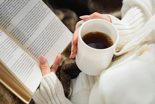 cropped view of woman reading book while holding cup of warm tea