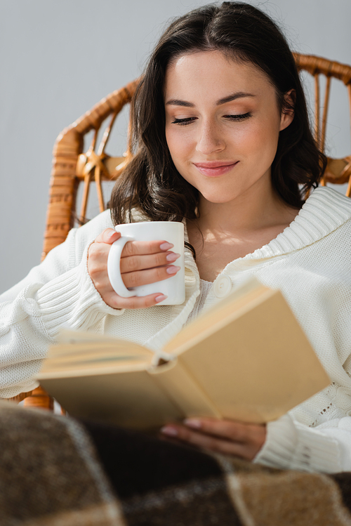 happy woman with cup of warm drink reading blurred book at home