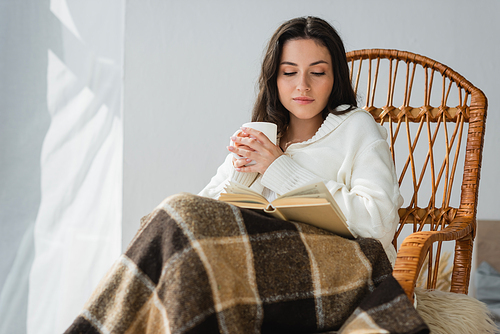 brunette woman sitting in wicker chair with cup of tea and reading novel