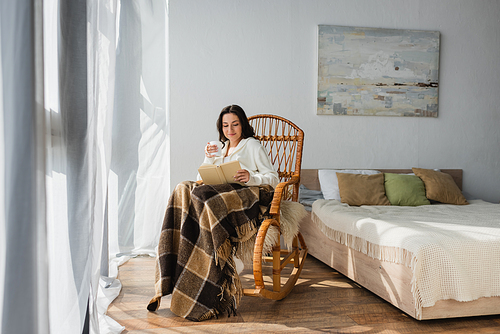 brunette woman reading book in bedroom while sitting in rocking chair under plaid blanket