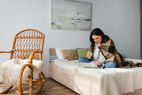 cheerful woman using laptop on bed near wicker chair