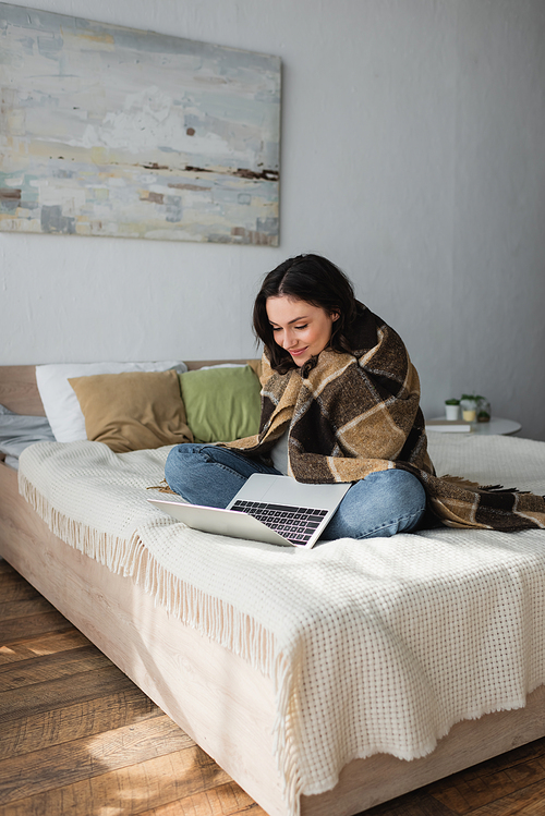 smiling woman sitting on bed under plaid blanket and looking at laptop