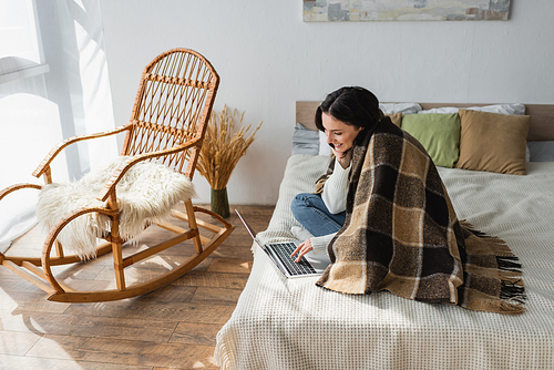 smiling woman sitting on bed near rocking chair and using laptop