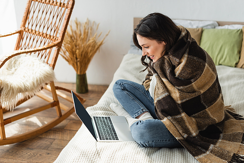 smiling woman looking at laptop with blank screen while sitting on bed under plaid blanket