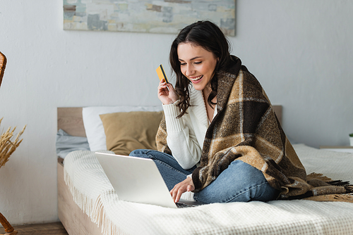 pleased woman sitting on bed under warm blanket while holding credit card near laptop