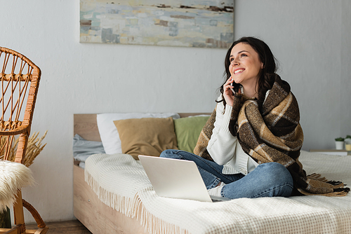 joyful woman sitting on bed near laptop and talking on mobile phone