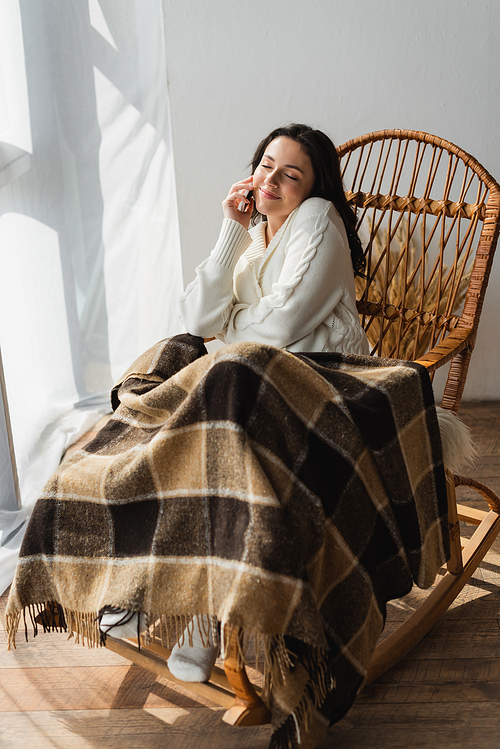 pleased woman sitting in rocking chair under warm blanket and talking on smartphone with closed eyes
