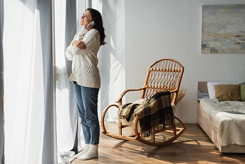 full length view of woman in jeans and white cardigan calling on smartphone near window and wicker chair