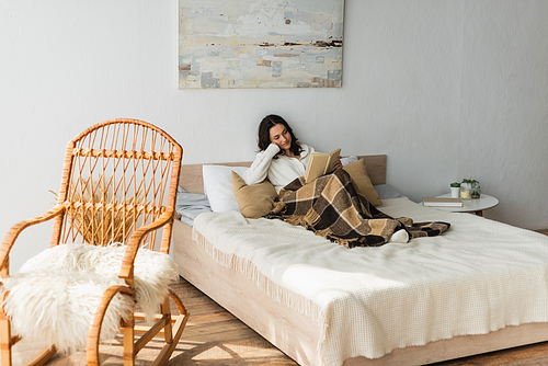 brunette woman reading book in bed under plaid blanket near wicker chair