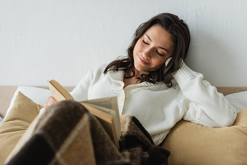 smiling woman in white cardigan reading book in bed under checkered blanket