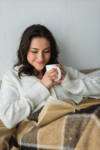 happy woman in warm cardigan drinking warm tea and reading book in bed