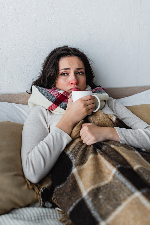 sick and displeased woman lying in bed with cup of warm tea under plaid blanket