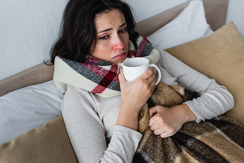 ill and upset woman lying under checkered blanket with cup of warm beverage