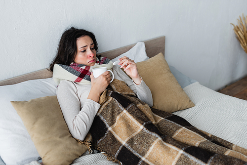 sick woman frowning while lying in bed and looking at thermometer