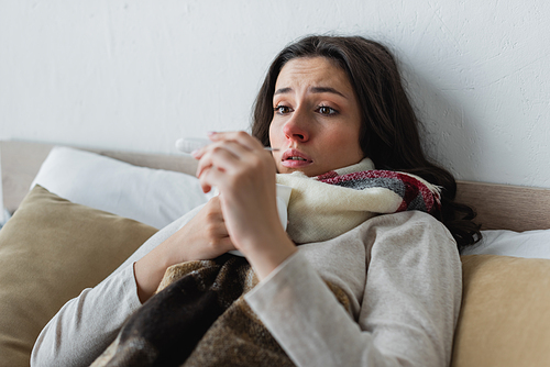 shocked and upset woman lying under warm blanket and looking at thermometer
