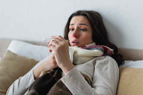 shocked woman looking at thermometer while lying in bed under plaid blanket