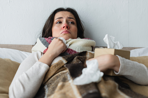 upset woman lying under plaid blanket near crumpled paper napkins, blurred foreground