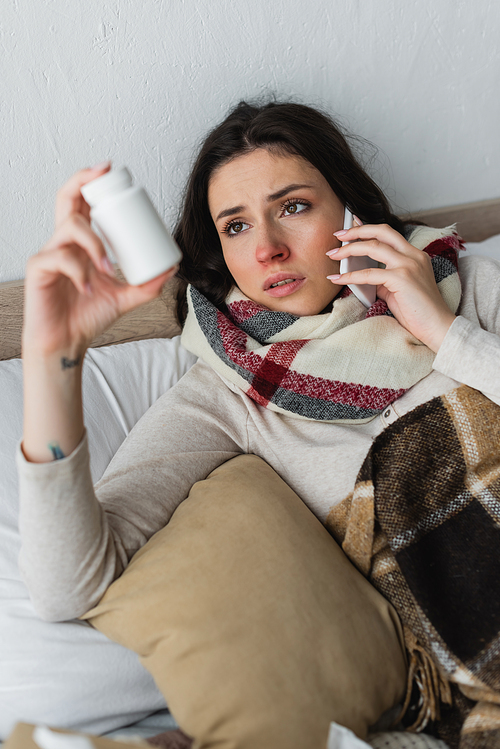 sick woman with pills container talking on mobile phone while lying under plaid blanket