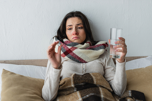 sick and sad woman holding glass of water and pill while lying under plaid blanket