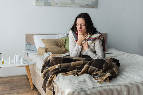 sick woman touching sore throat while sitting on bed under plaid blanket