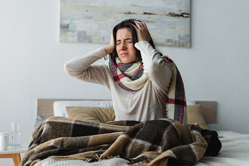 ill woman in warm scarf touching head while suffering from headache in bedroom
