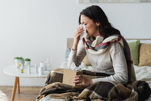 sick woman in warm scarf sitting under plaid blanket and sneezing in paper napkin