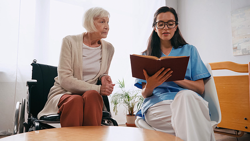 brunette geriatrician nurse reading book to senior woman in wheelchair