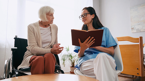 brunette geriatrician nurse in eyeglasses reading book to happy aged woman in wheelchair