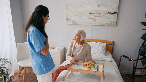 brunette nurse talking with aged patient near tray with breakfast on bed