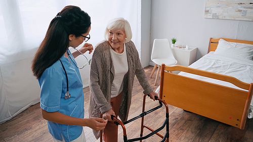 cheerful senior woman stepping with walkers near brunette nurse