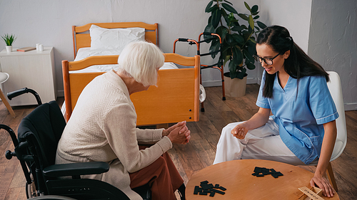 senior woman in wheelchair playing dominoes with happy nurse