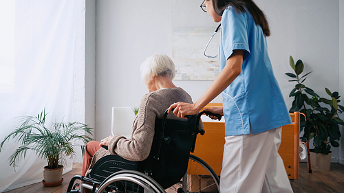 geriatric nurse in eyeglasses moving aged woman in wheelchair