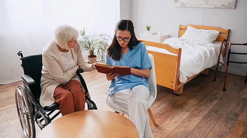 brunette geriatrician reading book to aged woman in wheelchair