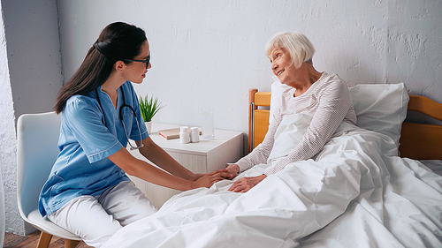 geriatric nurse in glasses talking with smiling aged woman in bed