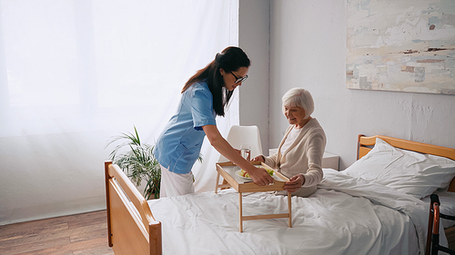brunette nurse carrying tray with breakfast to smiling aged patient