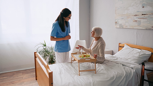 brunette nurse talking with cheerful senior patient near breakfast tray on bed
